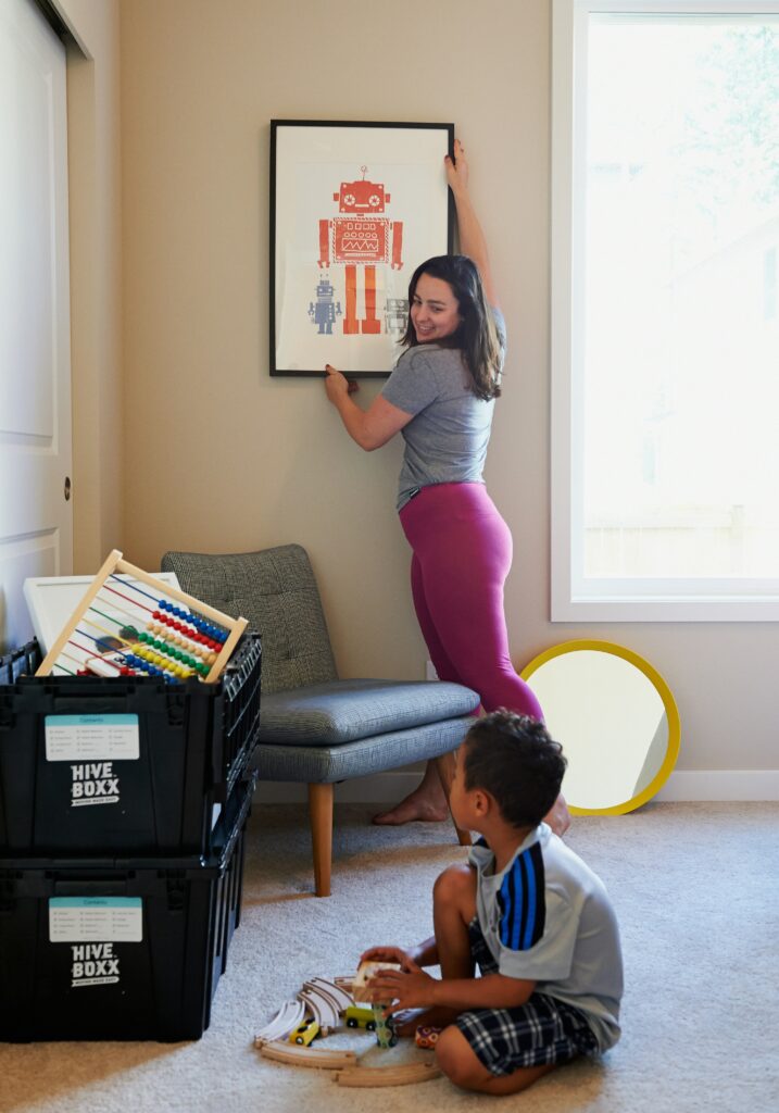 Happy young woman hanging a picture frame up in a new house whilst her child plays with wooden toys. 